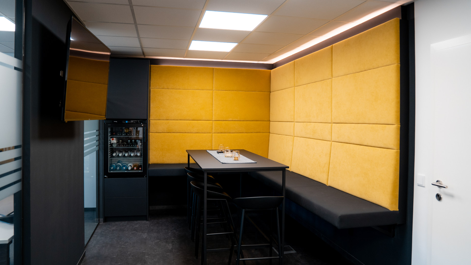 Moderner Büro-Loungebereich mit gelber Polsterwand, schwarzem Tisch und Bank, Glastrennwand, TV und Getränkekühlschrank.