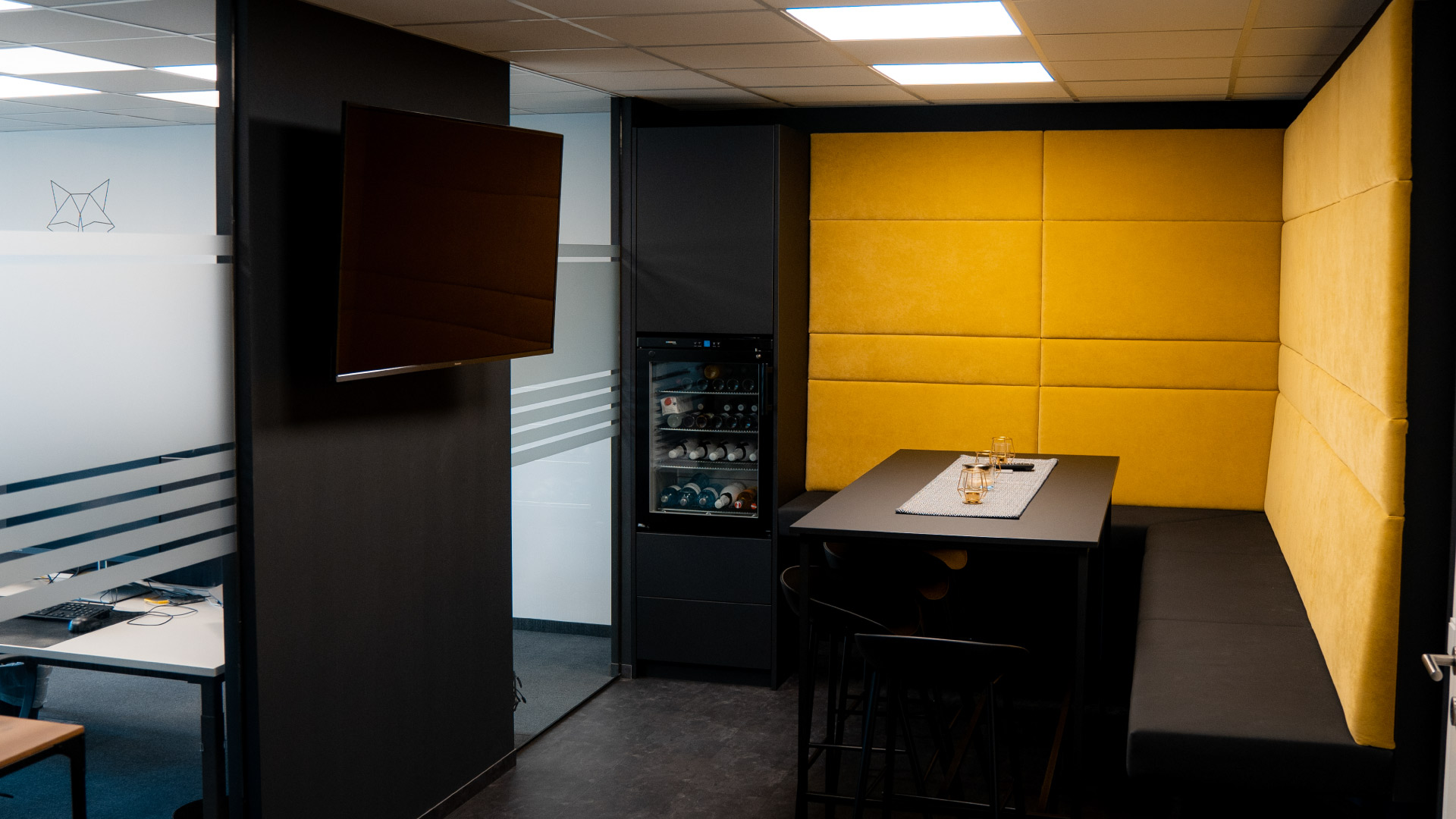 Moderner Büro-Loungebereich mit gelber Polsterwand, schwarzem Tisch und Bank, Glastrennwand, TV und Getränkekühlschrank.