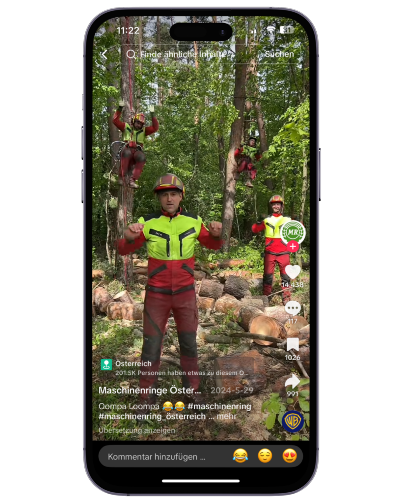 Ein TikTok-Video wird auf einem Smartphone angezeigt. Im Video sind mehrere Personen in rot-gelber Schutzkleidung und Helmen im Wald zu sehen. Zwei klettern auf Bäume, während vorne eine Person direkt in die Kamera blickt und Bewegungen macht. Im Hintergrund liegen gefällte Baumstämme.