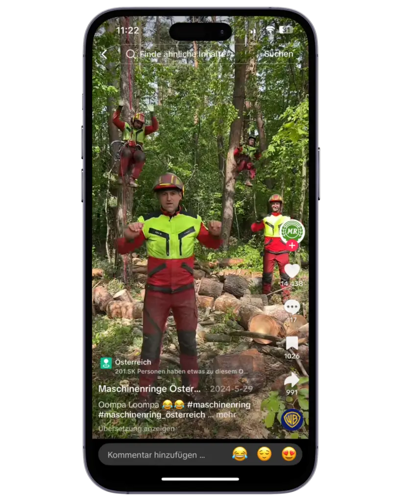Ein TikTok-Video wird auf einem Smartphone angezeigt. Im Video sind mehrere Personen in rot-gelber Schutzkleidung und Helmen im Wald zu sehen. Zwei klettern auf Bäume, während vorne eine Person direkt in die Kamera blickt und Bewegungen macht. Im Hintergrund liegen gefällte Baumstämme.