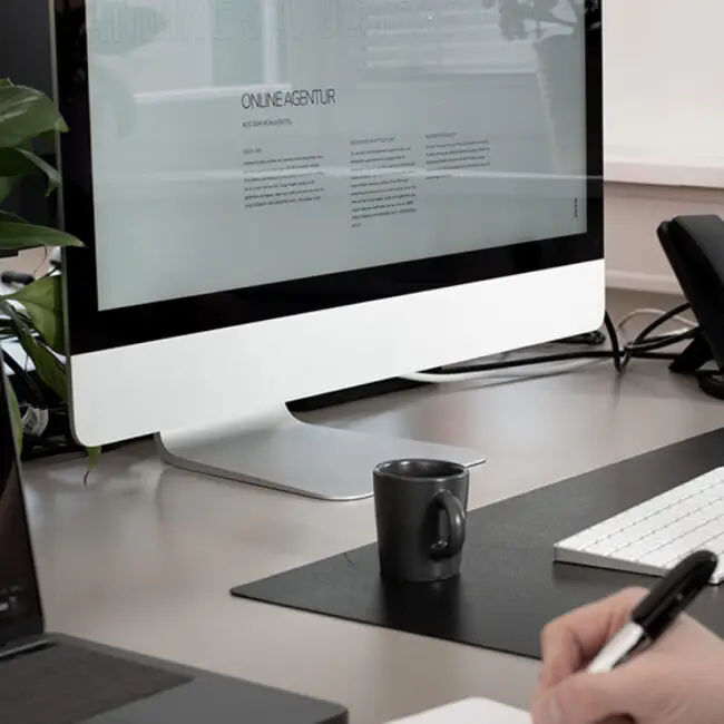 Schreibtisch mit iMac, Tastatur, Notizen und Kaffeetasse – Bildschirm zeigt Webseite einer Online-Agentur.