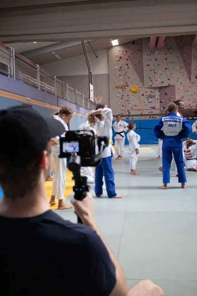 Eine Person filmt ein Judo-Training in einer Sporthalle; mehrere Judoka in weißen und blauen Anzügen üben auf der Matte, im Hintergrund ist eine Kletterwand zu sehen.